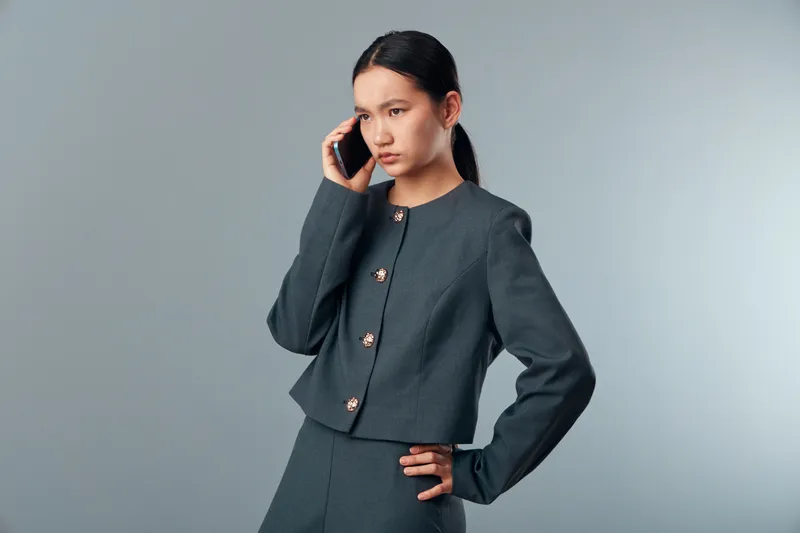 Young woman in dark suit talking on smartphone with serious expression standing isolated on gray background. Business professional, communication and focus in modern office style.