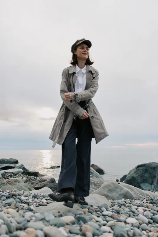 Young woman wearing a trench coat and cap stands on rocky shore near calm sea under cloudy sky. Casual style, autumn fashion, outdoor lifestyle, and nature concept in soft daylight.