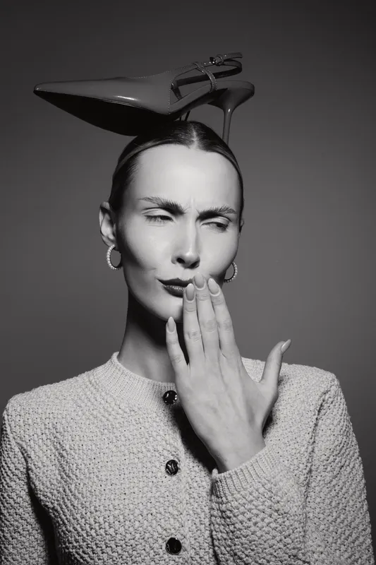 A fair-skinned Caucasian woman with slicked-back hair balances a high-heeled shoe on her head while covering her mouth with her hand. Concept of fashion, humor, and creativity in studio.