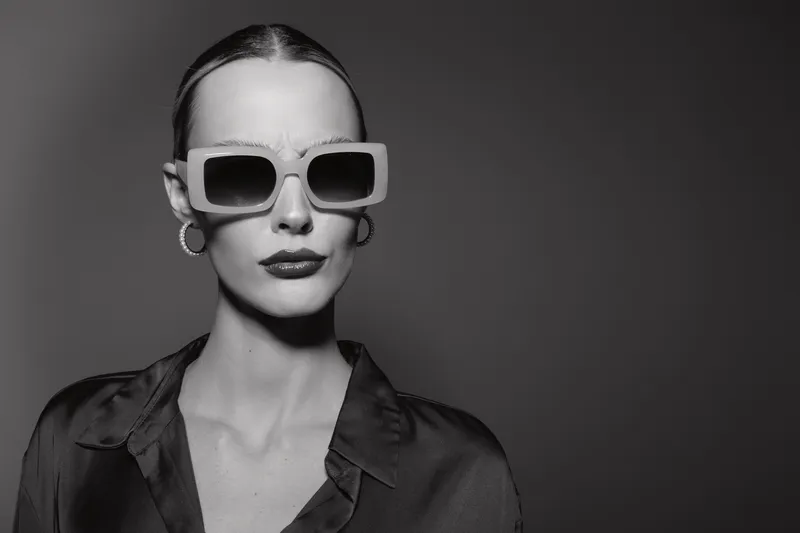 Stylish white woman with pale skin wears oversized sunglasses and hoop earrings. She poses confidently in a dark shirt against a plain grey background. Concept of fashion and attitude.
