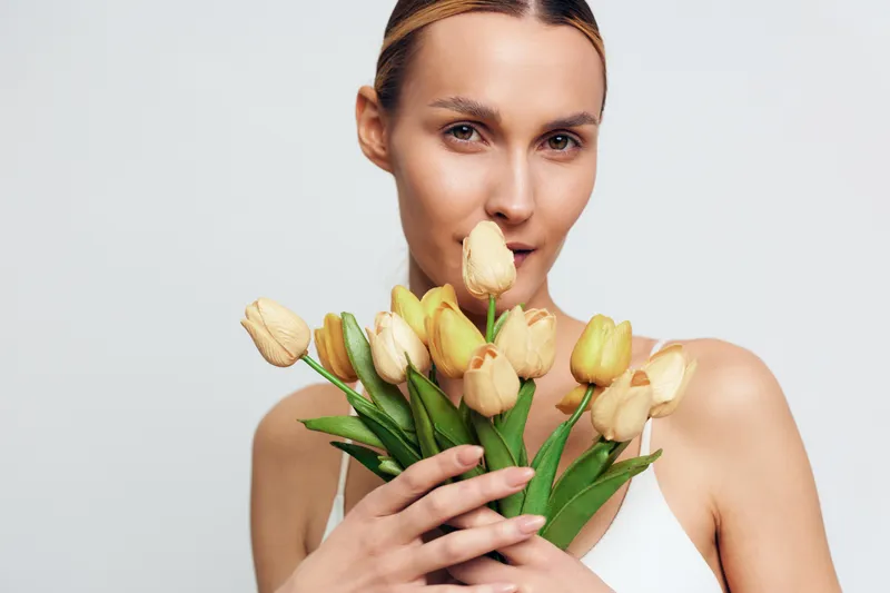 Young woman with blonde hair holds a bouquet of yellow tulips close to her face while looking softly at the camera against a plain light background.