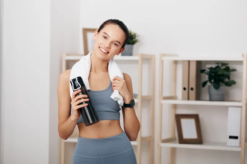 Smiling young woman in fitness apparel after workout, holding a water bottle with a towel over her shoulder, smartwatch visible, home gym interior with shelves and plant in soft light.
