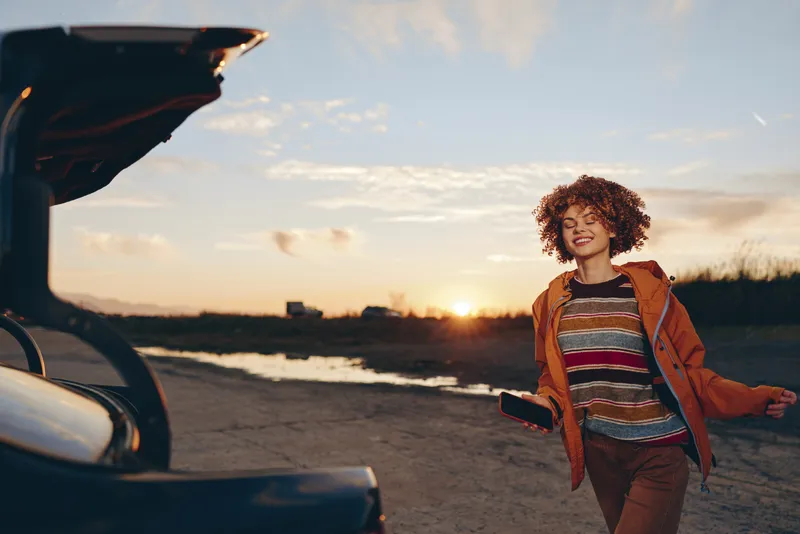 Smiling woman with curly hair by a car on a seaside road at sunset, travel lifestyle, warm sky, casual outfit, outdoor moment, soft natural light