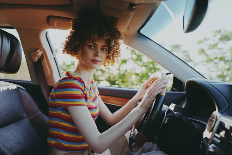 Young woman sits in a car, driving calmly, looking at the camera with a warm smile, striped shirt, curly hair, natural daylight, casual lifestyle portrait inside vehicle, relaxed travel moment