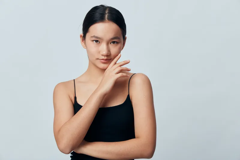 Young woman posing with hand near face wearing black sleeveless top against plain light background. Portrait with natural makeup and confident expression.