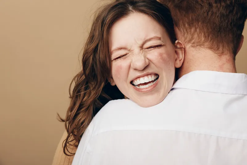 Happy woman with closed eyes and wide smile hugs man wearing white shirt. Close-up portrait on beige background. Concept of love, affection, joy, and emotional connection between couple.