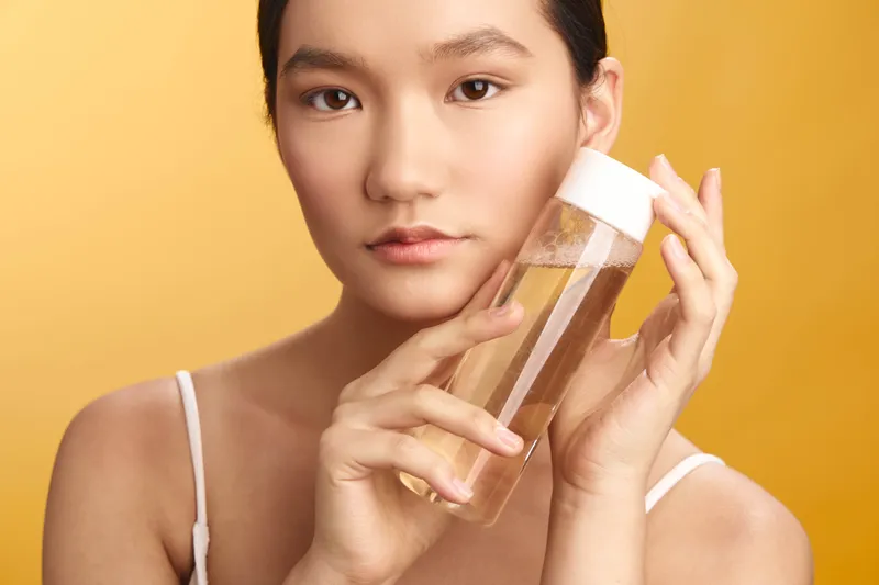 asian woman in a bright studio applies skincare product to her face, smiling softly and exuding confidence, creating a calm, modern beauty moment focused on self care.