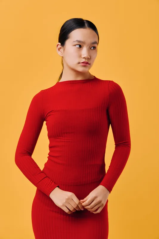 Young woman wearing red long sleeve dress standing in front of orange background looking sideways with hands clasped, posing with serious expression and neat hairstyle.