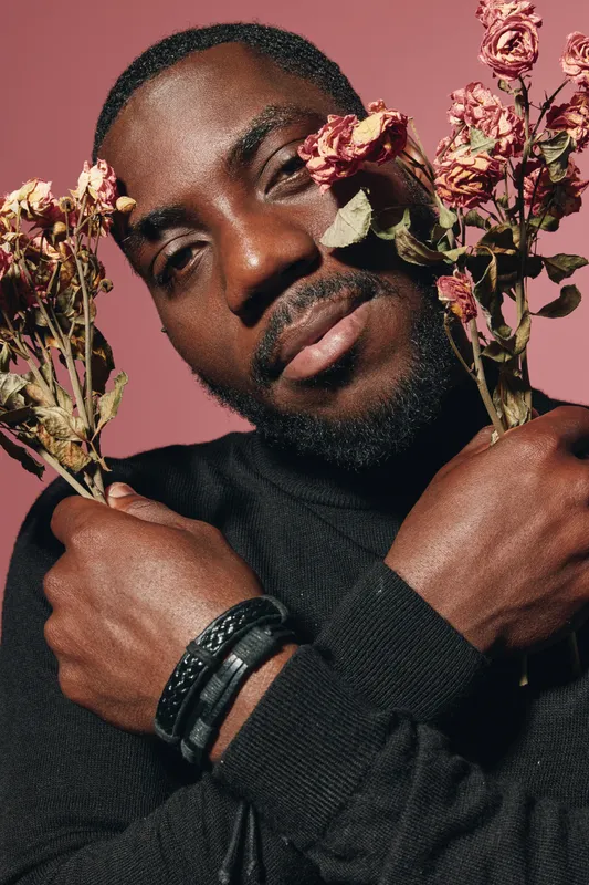 African man with dark skin holding dried roses in creative conceptual visual style. Portrait with closed eyes and black sweater on pink background for artistic expression.