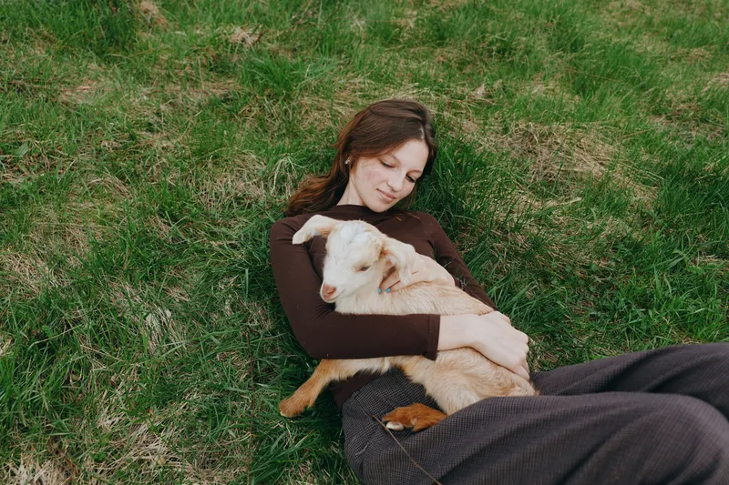 A woman rests on grassy ground, embracing a goat kid with a calm, tender expression. A wholesome lifestyle moment highlighting bonding, care, and rural life in natural light.