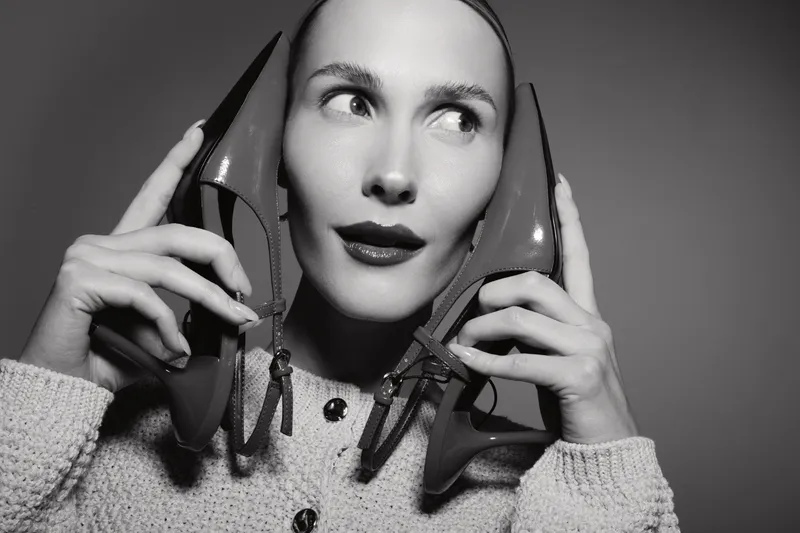 Portrait of a fair skinned white woman holding two high heel shoes near her ears, wearing a knitted sweater. Concept of fashion and creativity.
