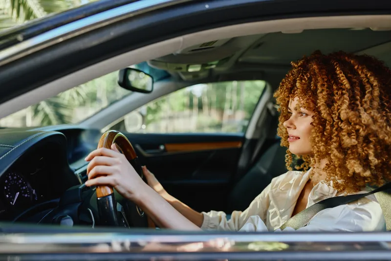 Curly woman driving car confidently through city with focus and bright sunlight highlighting her natural hair, wearing casual light-colored clothing inside modern vehicle interior.