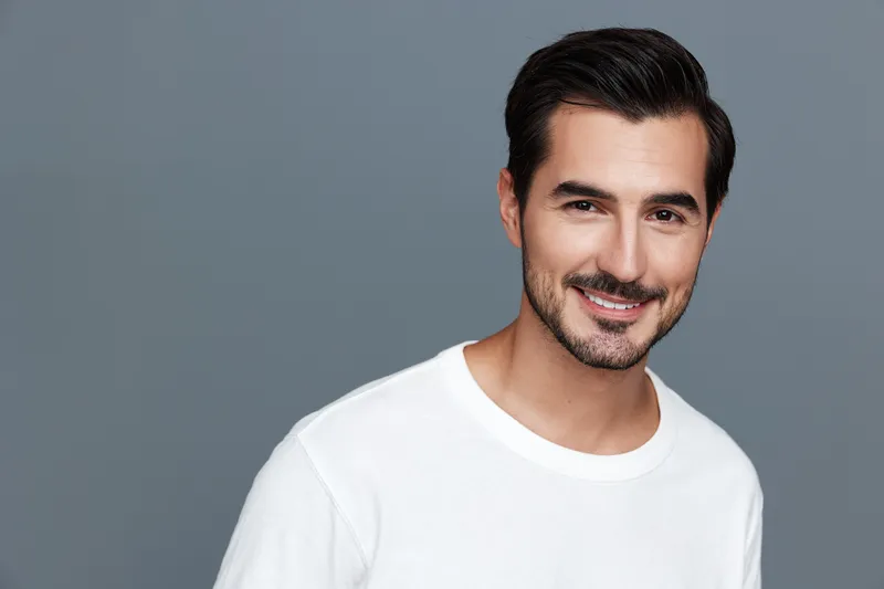 Smiling man in casual white t-shirt poses confidently against a grey background, showcasing a friendly expression and engaging demeanor. Portrait photography concept.