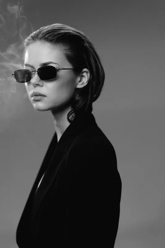 Black and white portrait of stylish woman wearing sunglasses and a dark blazer with smoke in the background.