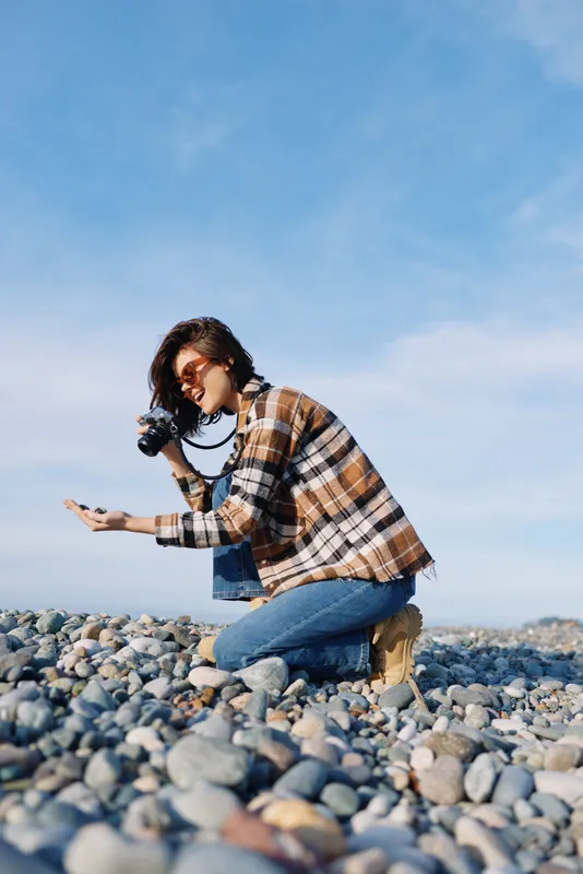 Photographer kneeling on a rocky beach with a camera, capturing candid outdoor moments in natural light, wearing a plaid shirt and jeans, focused on composition and textures with coastal light.