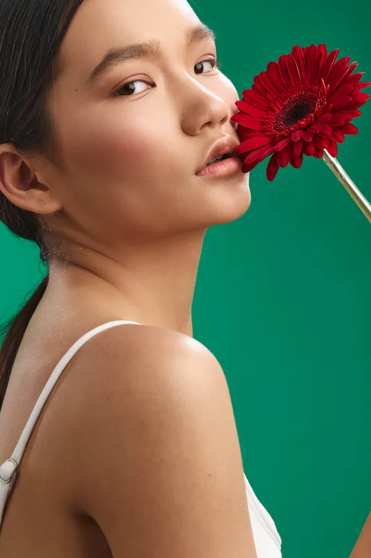 asian woman wears a white strap top and holds a red flower near her lips in a studio shot, set against a bold green background, creating a graceful, elegant and contemporary mood.