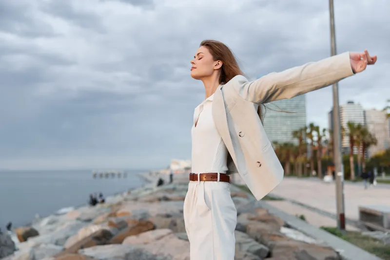 Confident woman in a business suit enjoying the seaside with open arms, embodying empowerment and freedom in a coastal city environment.
