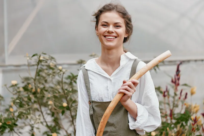 Smiling young woman in a greenhouse, wearing casual green overalls, holding a gardening tool, showcasing a connection to nature and sustainable gardening practices