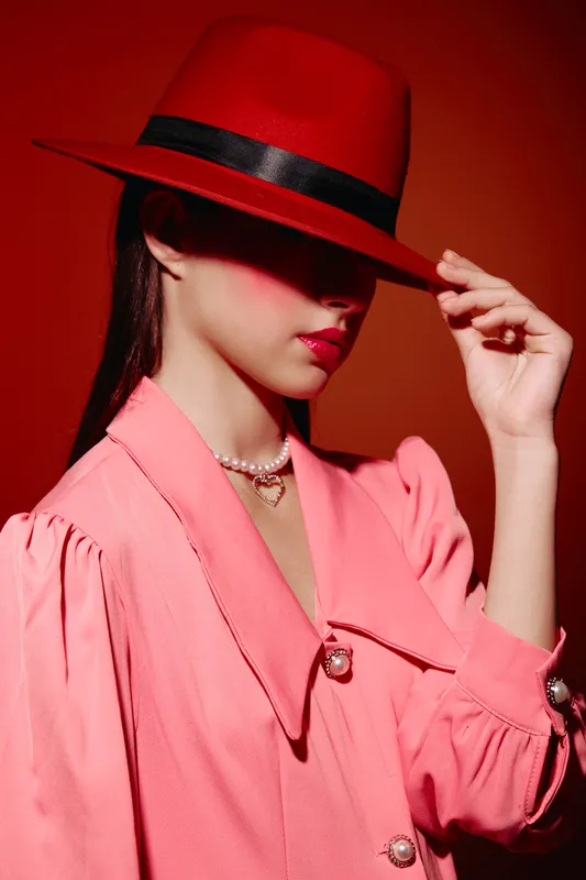 Stylish woman wearing a red hat with black ribbon and a pink tailored blazer, softly touching the brim, with a pearl necklace on red background beauty fashion concept.