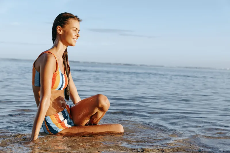 Woman in yoga pose on beach by ocean at sunrise, stretching for mindfulness and fitness, relaxation outdoors, soft natural light, summer near gentle waves and blue sky, healthy lifestyle