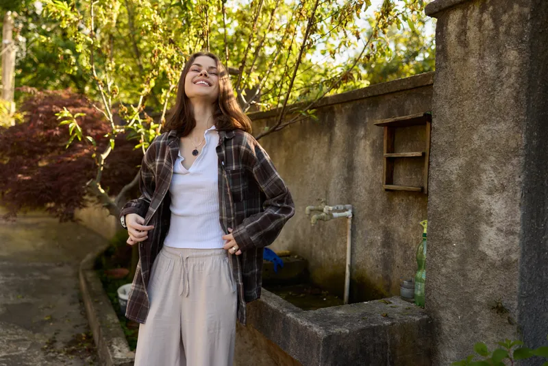 A joyful woman in casual clothing stands outdoors by a stone wall, smiling as sunlight filters through trees, capturing a light, everyday lifestyle moment