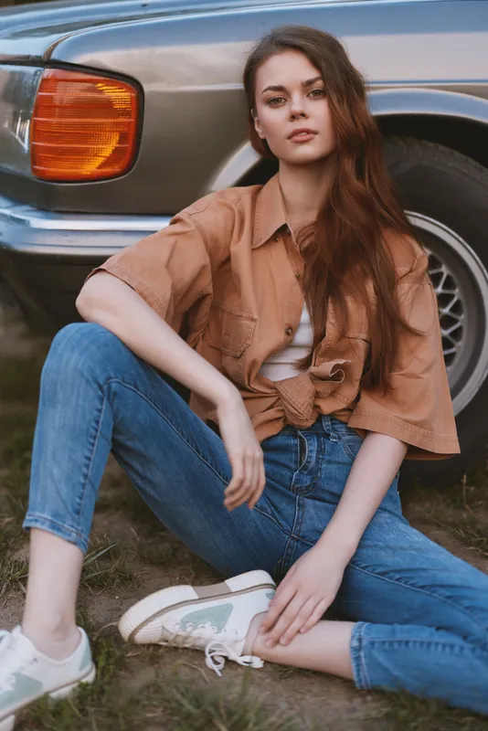 Stylish young woman in casual outfit sitting on the grass near a vintage car, showcasing a trendy summer look with relaxed denim and a soft color palette.
