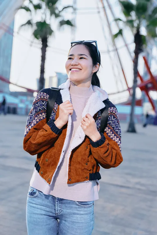 Woman with a warm smile wearing a patterned brown cream jacket and jeans, strolling along a sunny boardwalk with palm trees and urban backdrop, ready for travel, adventure, and casual street style