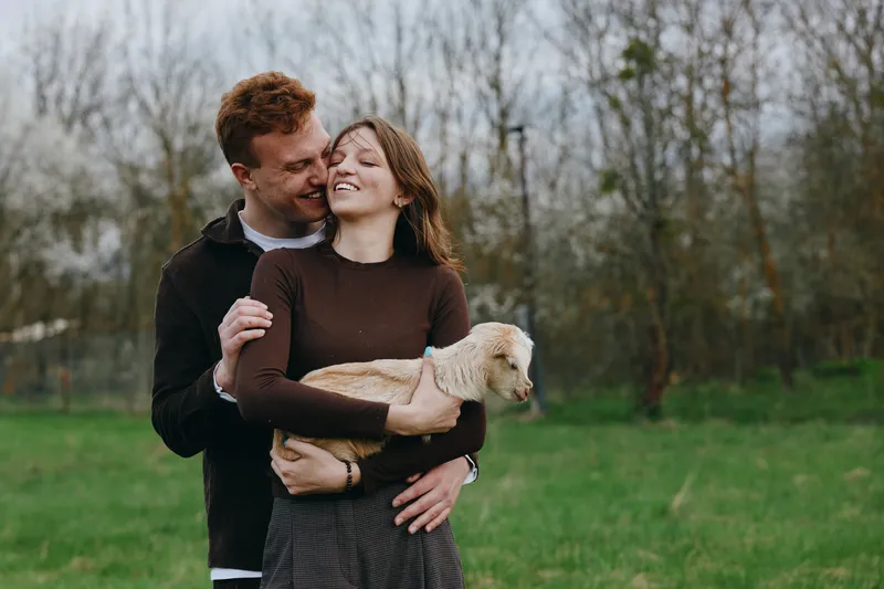 Happy couple outdoors in a grassy park share a warm moment while holding a light colored dog, smiling and affectionate, wearing cozy sweaters, enjoying a relaxed lifestyle and natural light.