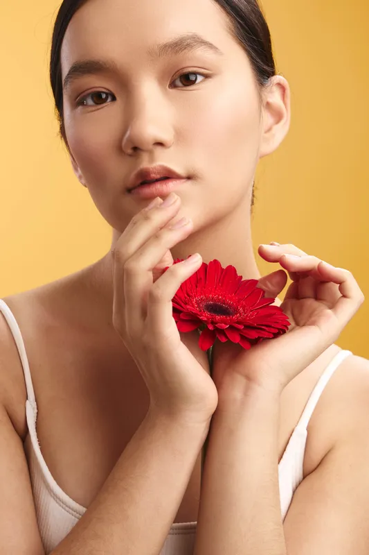 Asian woman holds a vibrant red flower near her face against a warm yellow backdrop, creating a calm studio portrait with elegant, natural light and subtle beauty.