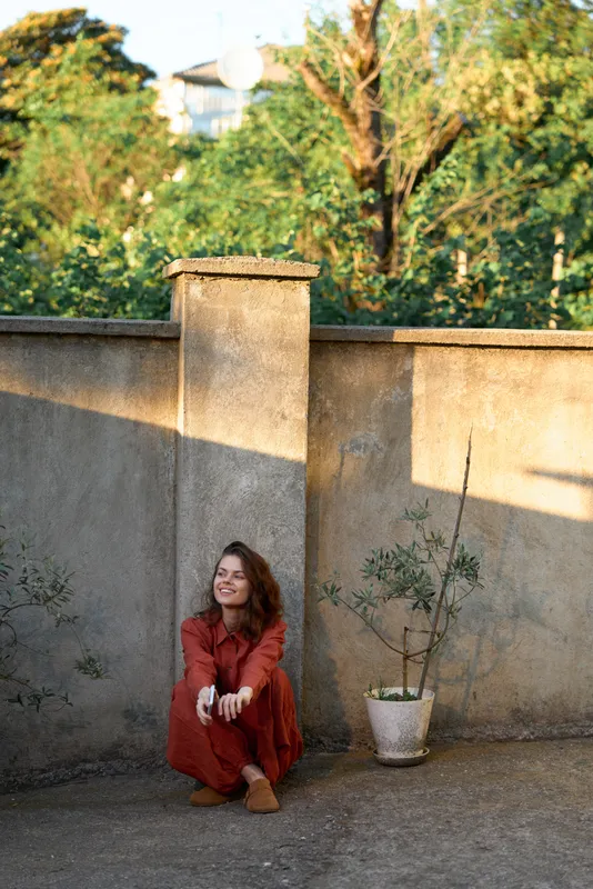 Sunlit woman in a red dress sits on the ground against a warm wall, smiling softly and relaxed in a calm outdoor moment