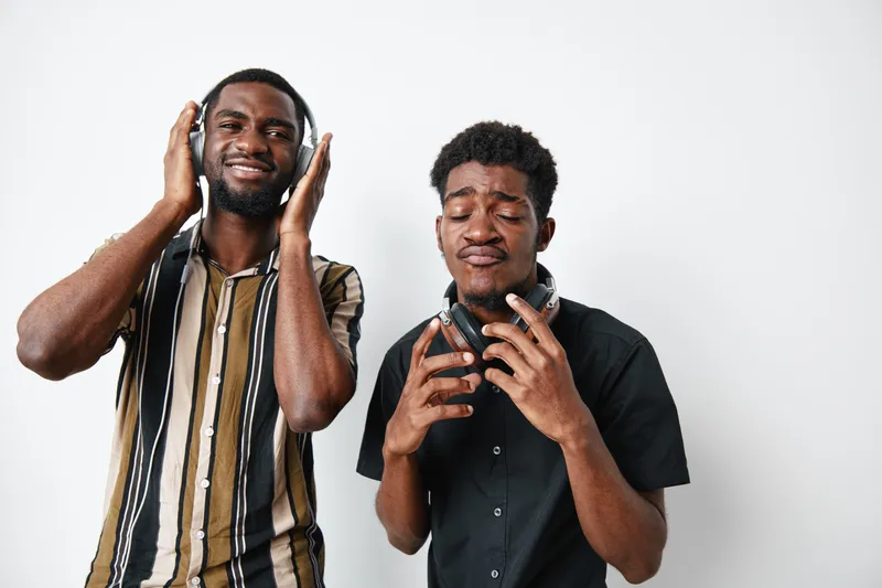 Two young black men with dark skin enjoying music with headphones, expressing emotions and having fun together in casual clothing on white background.