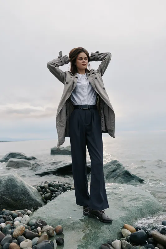 Young woman in stylish grey coat and white shirt stands on rocky shore by the sea with arms raised behind head. Overcast sky and waves create moody outdoor fashion concept.