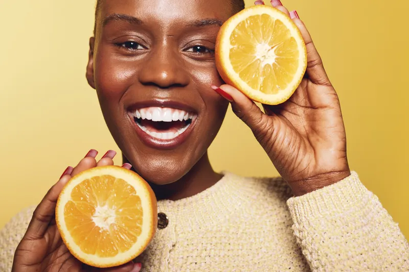 Happy smiling black woman with dark skin holds two halves of fresh orange fruit near face. Yellow background and cozy sweater create warm and joyful concept of health and natural beauty.