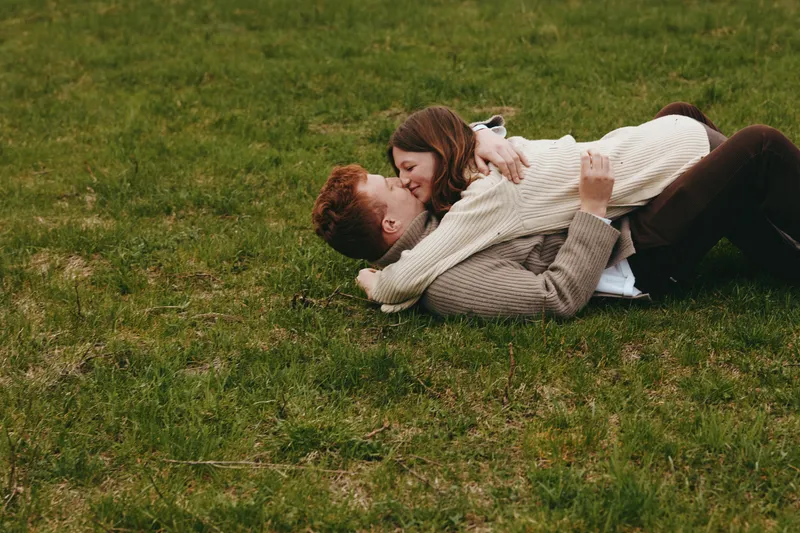 Romantic couple lying on grass outdoors, embracing in a sunny park, candid lifestyle moment with warm textures, natural light, and affectionate smiles, showcasing connection and peaceful togetherness