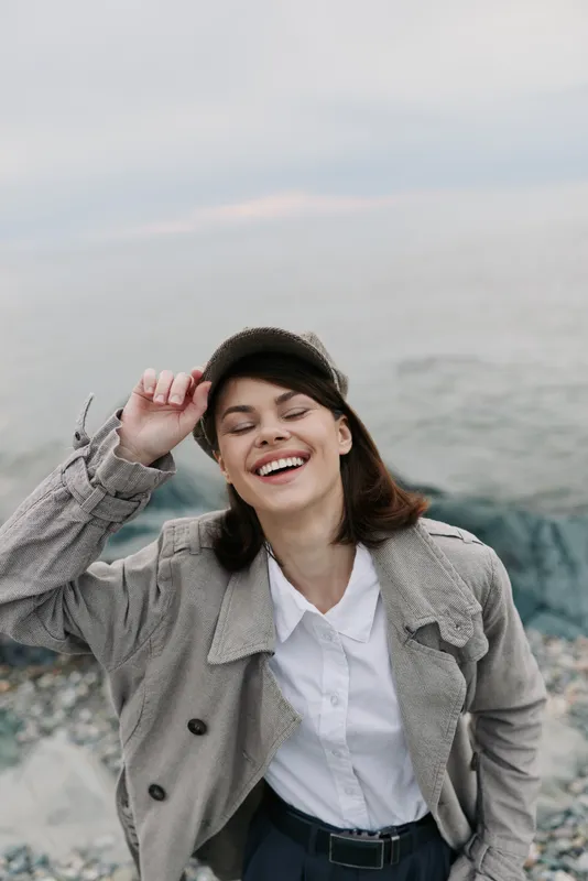 Happy young woman wearing a jacket and white shirt smiles with eyes closed near a rocky shore. Natural light highlights joyful mood and casual fashion by the sea.