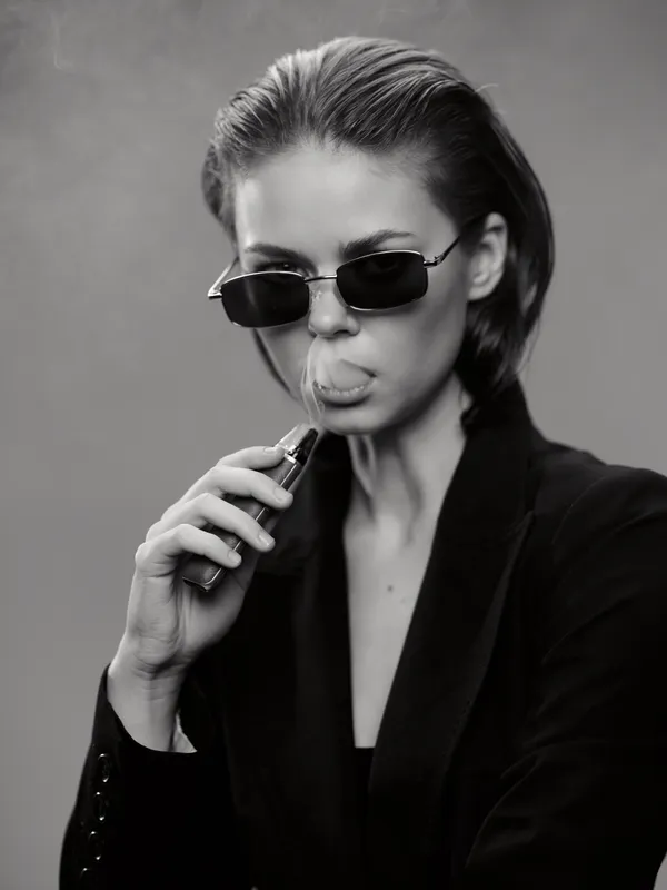 Black and white portrait of a stylish woman wearing sunglasses vaping with cool attitude and modern fashion vibes.