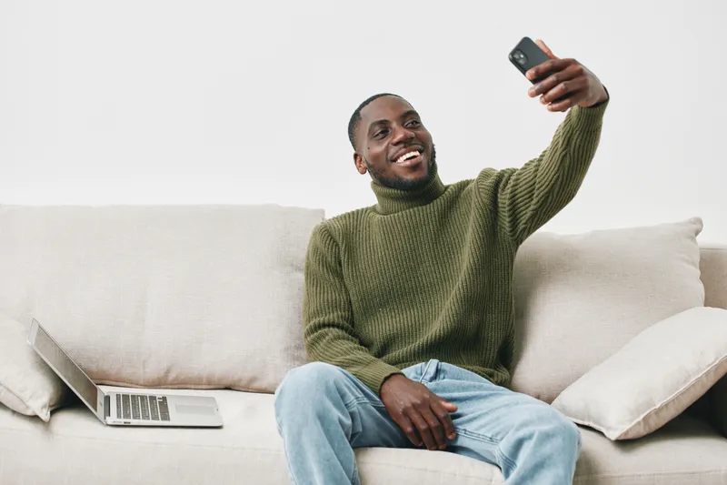 Happy African American man taking a selfie on a couch, wearing a green sweater, promoting self confidence and positivity in a cozy home environment