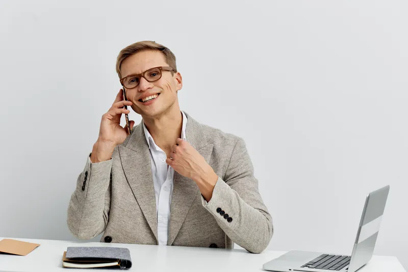 Smiling man in business casual attire talking on mobile phone while sitting at a desk with a laptop and notebook, isolated on plain white background, studio shot. Business communication concept