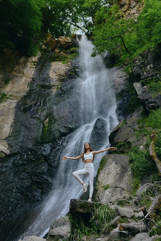 woman practicing yoga near waterfall in lush green forest wearing white activewear, balancing on one leg on rock, serene natural environment