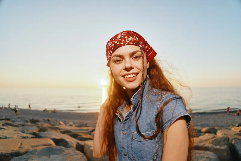 Lifestyle woman wearing modern boho-western denim street style with a red bandana, smiling by the beach at sunset in warm film color tones, embodying freedom and joy.