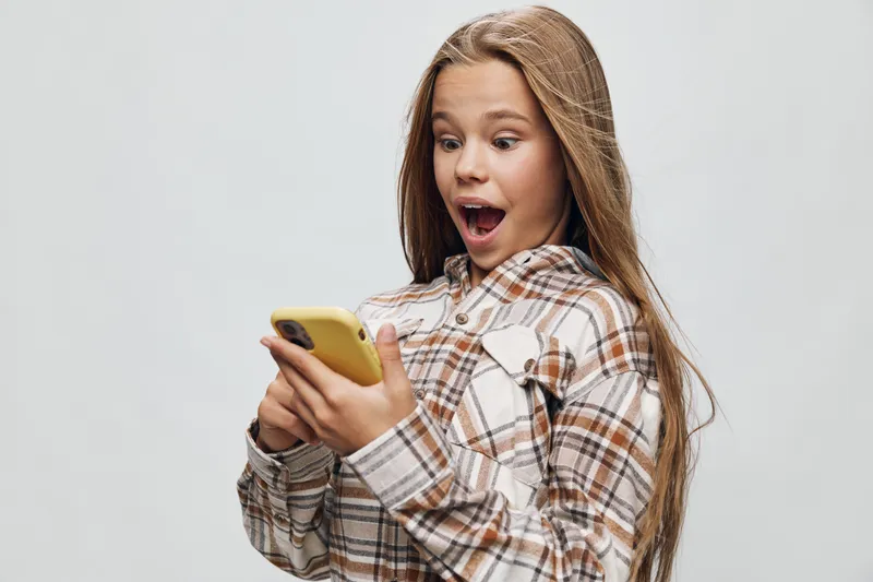 Girl with long hair wearing plaid shirt looking surprised while holding yellow smartphone with both hands on gray background. Child expressing emotion and digital communication concept.