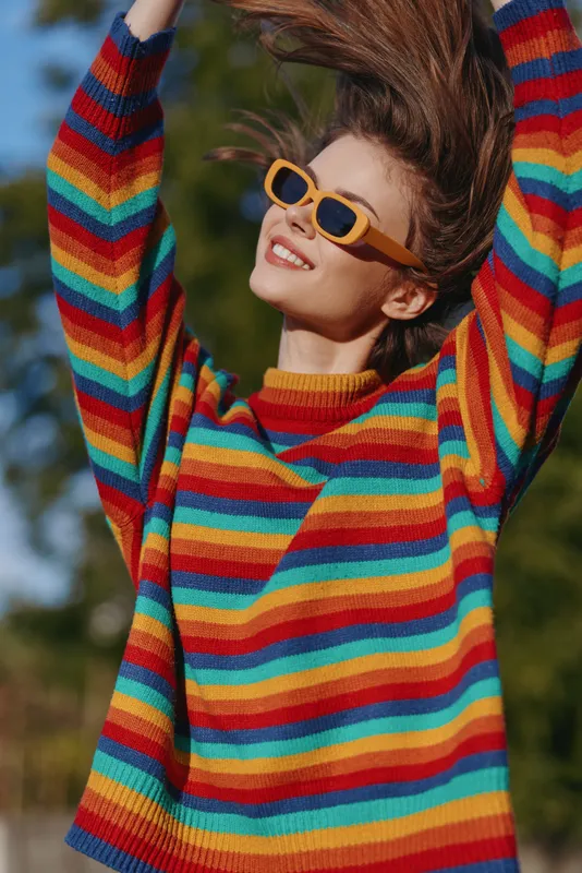 Woman in a rainbow sweater wearing yellow sunglasses, smiling outdoors in warm sunlight. Youth model with casual outfit, hair flying while posing joyful, colorful fashion lifestyle portrait.