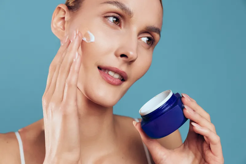 A young woman applies moisturizer cream on her face holding a blue jar, showing skincare routine and healthy skin care concept with soft natural light on blue background.