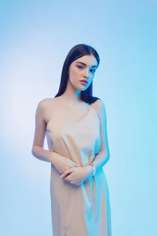 Young woman beauty fashion model with long dark hair poses in a pale dress against a soft blue background. Studio portrait with elegant and minimalist style concept.