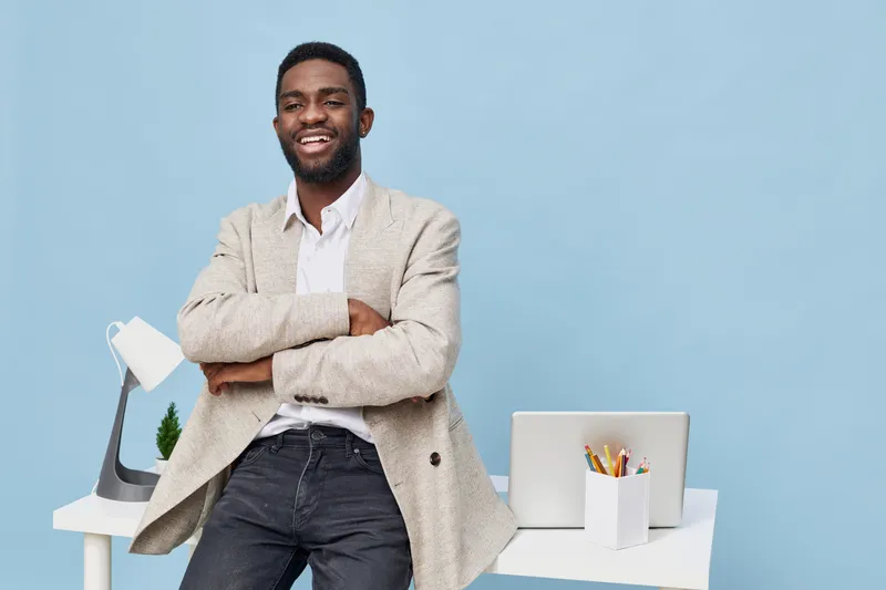 Confident smiling man with dark skin, casual beige blazer, white shirt, standing in modern office with pastel blue wall, laptop, and stationery on desk, creating an inviting professional atmosphere. P