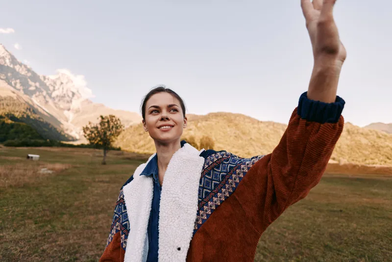 Woman portrait in mountains and nature smiling outdoors, wearing a warm jacket and casual layers. Landscape scenery with meadow and sky, young female enjoying hiking, freedom and relaxation.