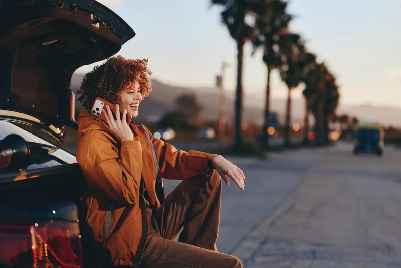 Woman in winter jacket opens car door on city street, smiles while waving a hand, travel lifestyle moment in warm sunset light, urban road background, candid photo for lifestyle