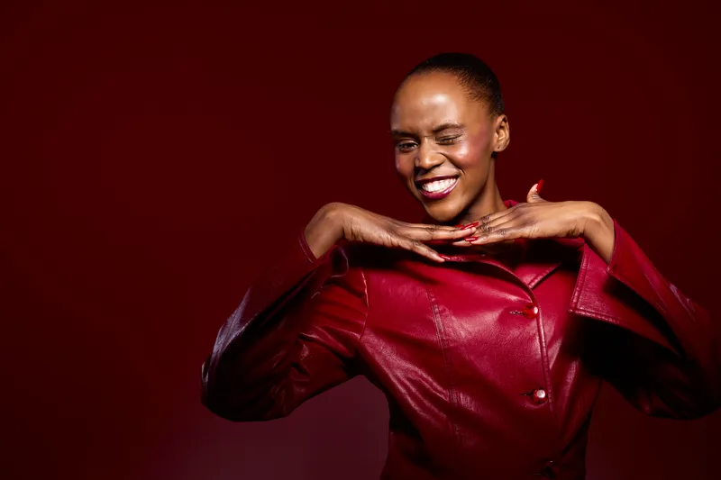 Smiling African woman with dark skin wearing red leather jacket poses happily against red background. Concept of fashion, confidence, beauty, and joyful expression in studio lighting.