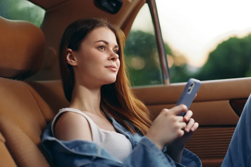 Young woman enjoying her time in a car, using a smartphone, wearing casual clothing, showcasing a relaxed emotion and warm ambient light.