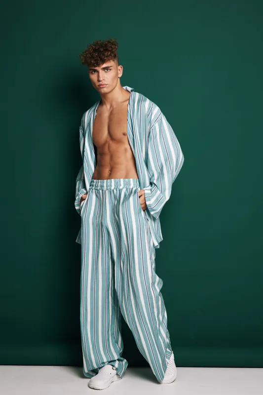 Young man posing in oversized striped pajamas with opened shirt and loose pants on green background, casual style and relaxed look with modern haircut and hands in pockets.